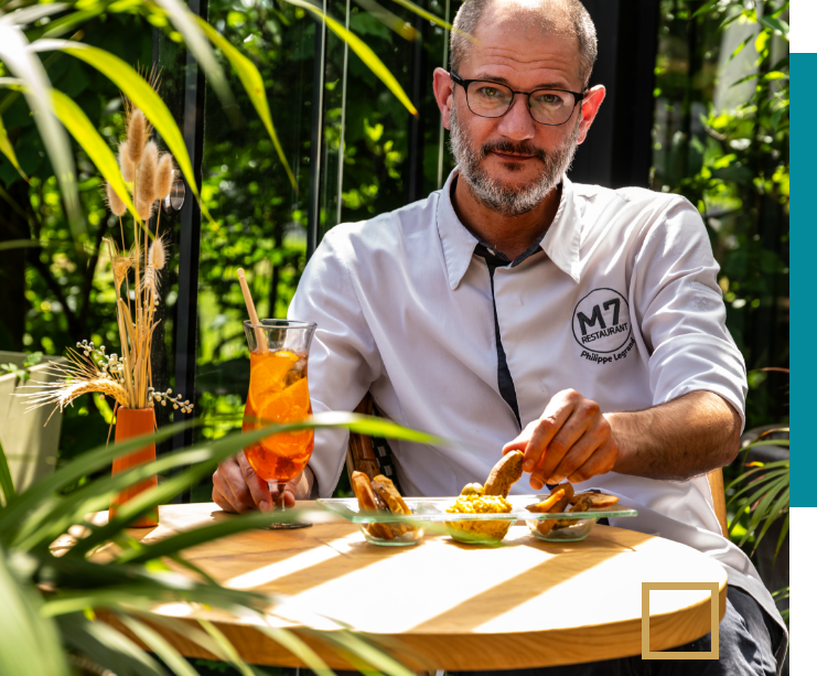 Chef du restaurant M7 assis en terrasse, devant une boisson estivale et des amuse-bouches.